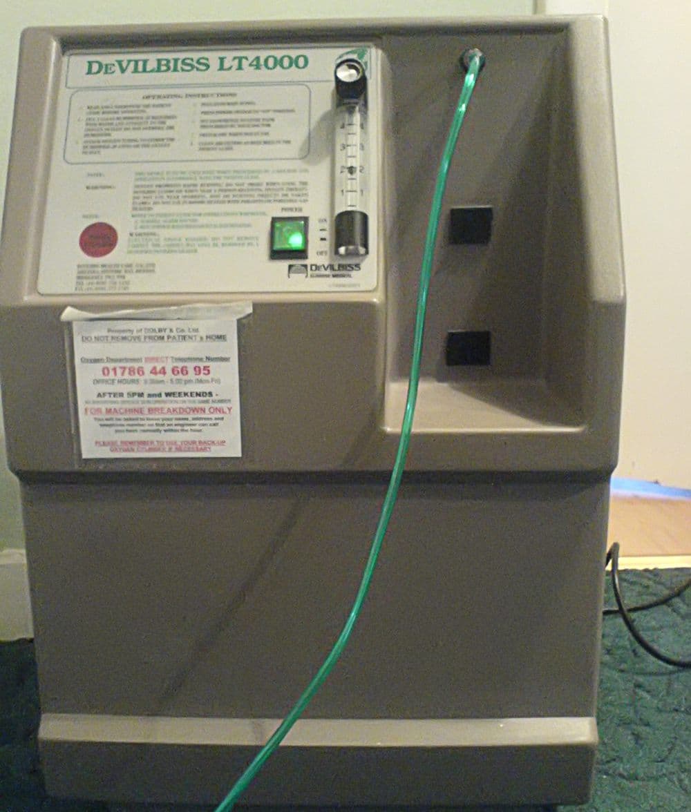 A beige DeVilbiss LT4000 oxygen concentrator with green tubing and a printed sticker that says it is for machine breakdown calls only