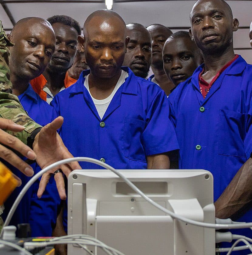 Biomedical technicians in blue work coats gather around a patient monitor during a training session